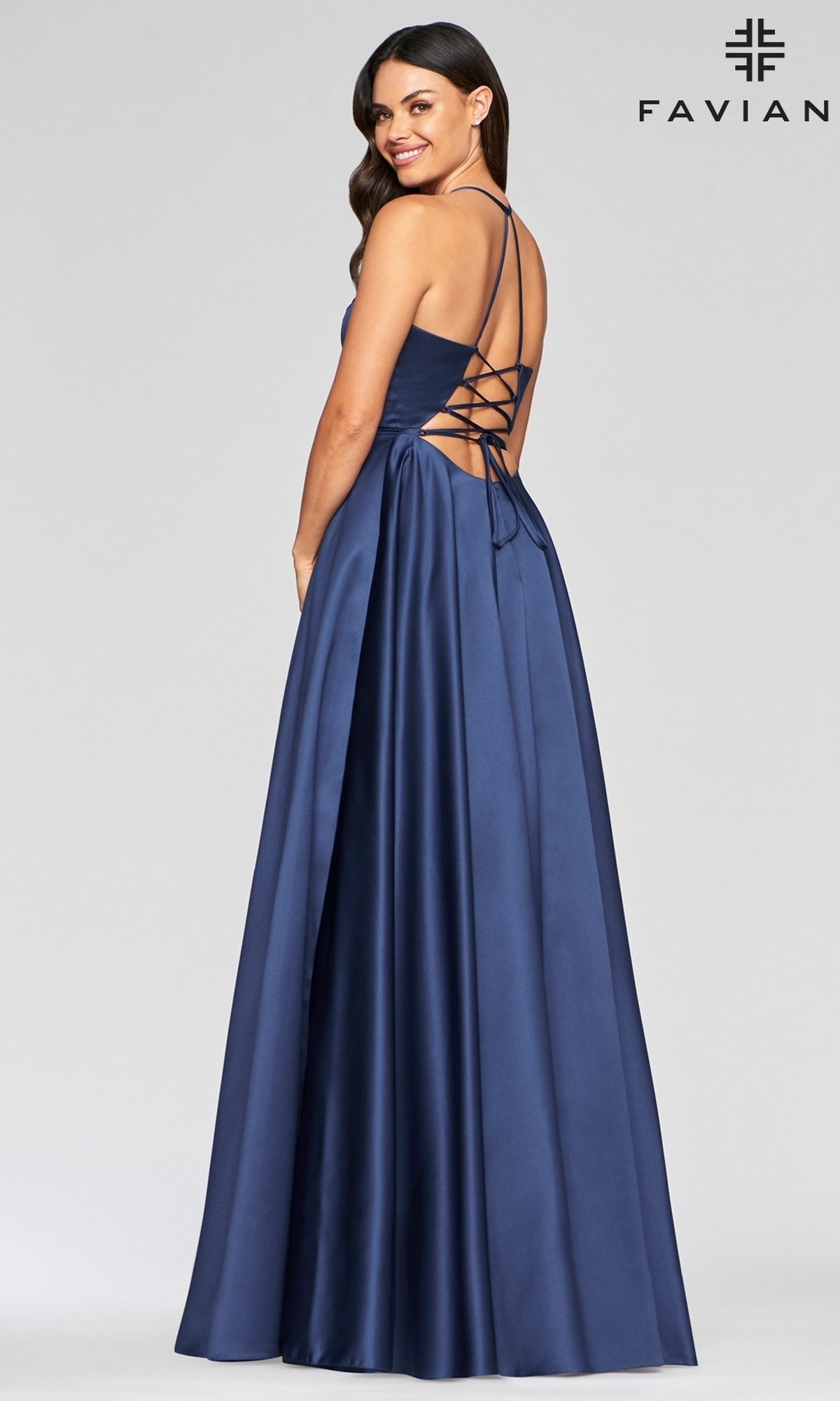 A woman smiles while standing sideways in a navy Corset-Back A-Line Long Satin Prom Dress by Faviana, featuring a satin finish, lace-up back, and spaghetti straps. The